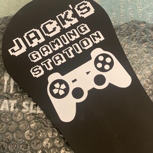 May include: Black gaming station sign with white text reading "JACK'S GAMING STATION" and a white controller graphic. The sign is paddle-shaped.