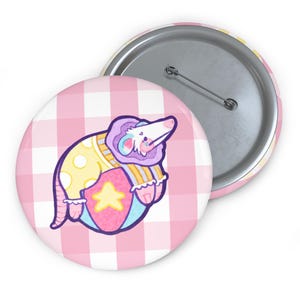 May include: Two round buttons. One button features a cartoon rat in a yellow polka-dot outfit, resting on a colorful ball with a star. The background is a pink and white checkered pattern. The second button is silver with a pin.