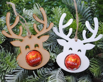 May include: Two reindeer-shaped Christmas ornaments. One is light brown, the other white. Each has a Lindor chocolate ball as a nose and a twine loop for hanging. Set against a backdrop of evergreen branches.