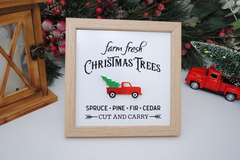 Puede incluir: Un cartel enmarcado con el texto "farm fresh CHRISTMAS TREES" y un cami&oacute;n de juguete rojo que lleva un &aacute;rbol de Navidad verde. El cartel tambi&eacute;n incluye el texto "SPRUCE - PINE - FIR - CEDAR - CUT AND CARRY - "