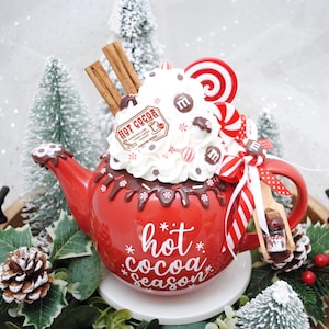 May include: A red ceramic teapot decorated with whipped cream, chocolate drizzle, and candies. The teapot has the words "hot cocoa season" written on it. Cinnamon sticks and a small wooden spoon are included.