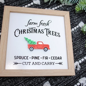 Puede incluir: Un cartel enmarcado con el texto "farm fresh CHRISTMAS TREES" y un cami&oacute;n rojo con un &aacute;rbol de Navidad en la caja. El cartel tambi&eacute;n dice "SPRUCE - PINE - FIR - CEDAR" y "CUT AND CARRY".