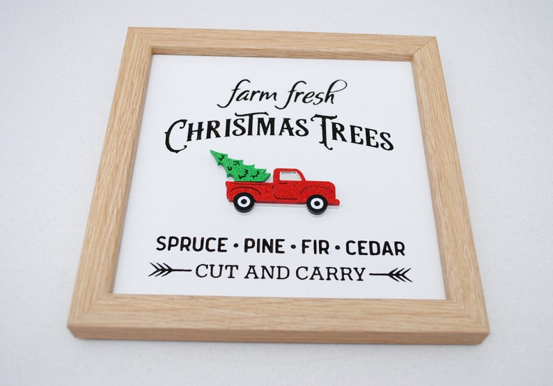 Puede incluir: Un cartel enmarcado con el texto "farm fresh CHRISTMAS TREES" y un cami&oacute;n rojo con un &aacute;rbol de Navidad en la parte trasera. El cartel tambi&eacute;n dice "SPRUCE - PINE - FIR - CEDAR" y "CUT AND CARRY" con flechas.