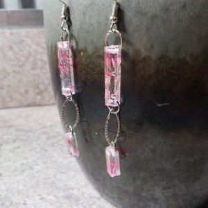 May include: A pair of silver dangle earrings with pink and white resin rectangular charms. The charms have a floral pattern.