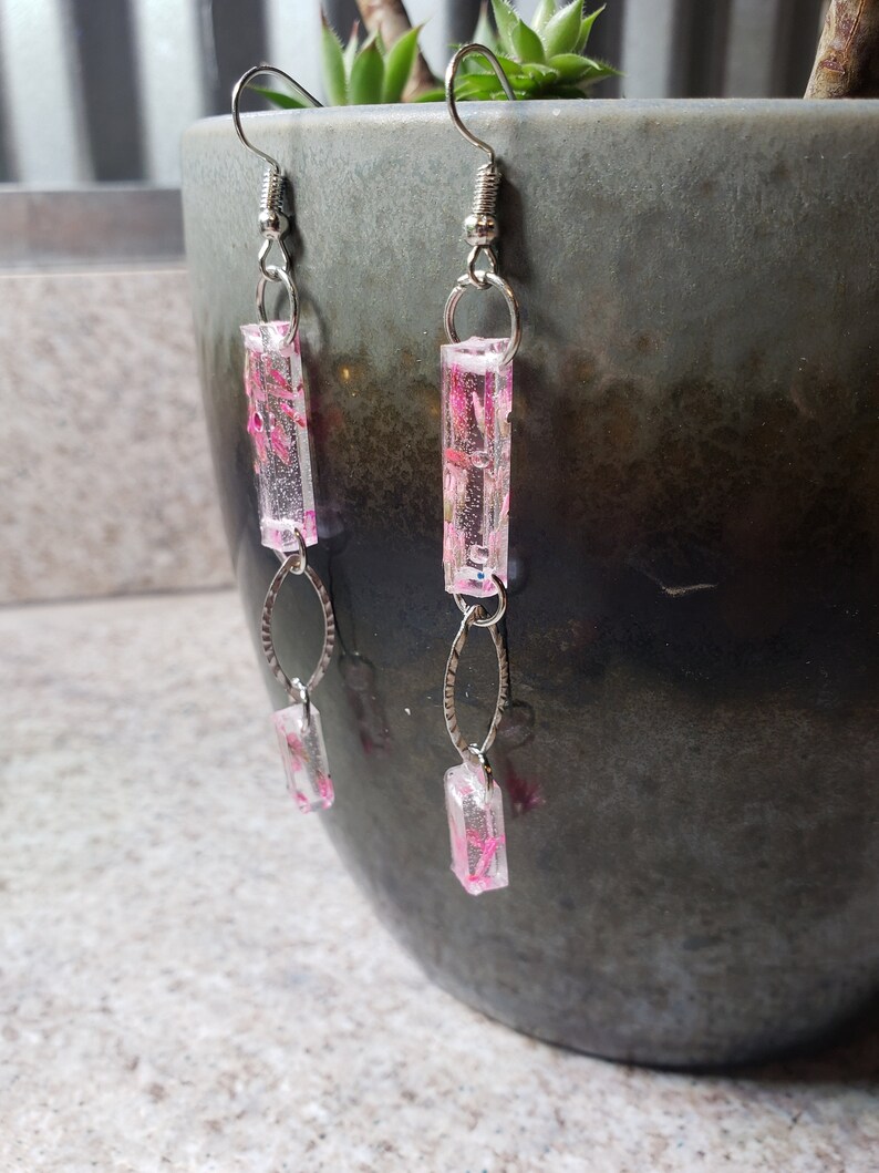 K&ouml;nnte beinhalten: Ein Paar silberne Ohrringe mit pinkfarbenen und wei&szlig;en rechteckigen Anh&auml;ngern aus Harz. Die Anh&auml;nger haben ein Blumenmuster.