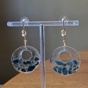 May include: A pair of gold hoop earrings with a clear resin circle containing small green and blue pieces of stone.