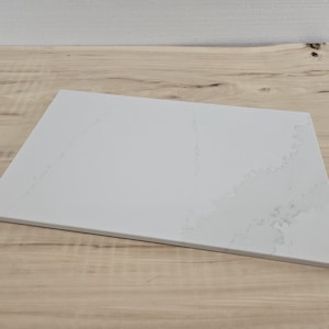 May include: A rectangular, white countertop with subtle gray veining. The countertop is set on a light-colored wooden surface. The countertop's clean lines and neutral color palette suggest a modern aesthetic, suitable for kitchen or bathroom design.