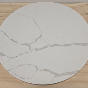 May include: A round, white countertop with gray veining. The countertop is made of a solid material and has a smooth surface. The countertop is set on a light-colored wooden surface.