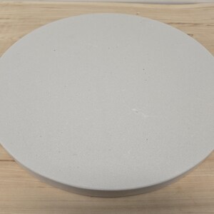 May include: A circular, light grey stone tray with a smooth, flat surface and a subtle speckled texture. The object features a clean, rounded edge and a solid, uniform appearance, presenting a simple, functional design for display or organisation.