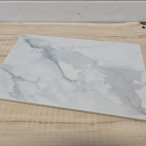 May include: A rectangular serving board featuring a white surface with prominent grey marble veining patterns. The board has a smooth, flat top and straight edges, showcasing a classic stone-like appearance with a high-contrast grey and white finish.