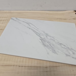 May include: A rectangular serving board featuring a white surface with prominent grey veining that mimics the appearance of natural marble. The board has a smooth, flat top surface and straight edges, providing a clean and simple design.