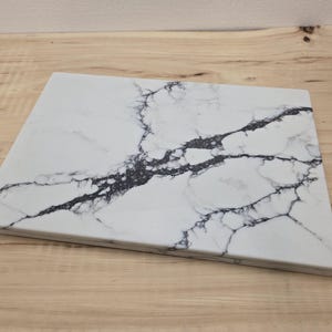 May include: A rectangular white marble chopping board with dark grey veining. The board is set on a light wood surface. The marble pattern creates a striking contrast against the white background, suitable for kitchen use.