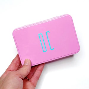 Puede incluir: Una lata rectangular rosa con las letras "DC" en azul turquesa.