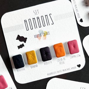 Pode incluir: Um conjunto de tintas aquarela feitas à mão em uma variedade de cores, incluindo preto, amarelo, roxo, laranja e rosa. O conjunto é rotulado como "Bonbons" e inclui o texto "Douceur Colorée".