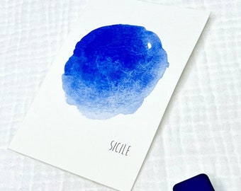 May include: A white card with a blue watercolor design and the word "Sicile" printed in black. A small, square, blue paint block is next to the card. The background is a white, textured fabric.