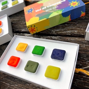 Pode incluir: Um conjunto de seis pastilhas quadradas de tinta aguarela numa caixa de cartão branca. As cores incluem laranja, verde, azul escuro, vermelho vivo, verde azeitona e amarelo. A tampa apresenta um padrão floral colorido com o texto Nid de campagne.
