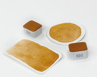 May include: A collection of miniature food items, including a pancake on a white plate, a rectangular piece of toast, and two square blocks with the text "ODO" on them. The food items are various shades of brown.