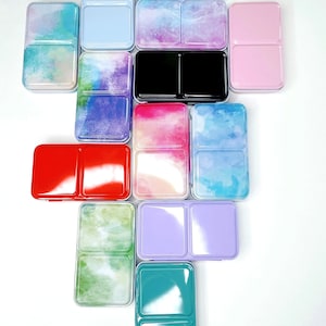 Peut inclure: Un ensemble de 12 godets de peinture aquarelle de différentes couleurs et motifs. Les godets sont disposés en forme de pyramide. Les godets sont en métal et ont une finition brillante. Les motifs sur les godets comprennent des lavis d'aquarelle, des motifs abstraits et des couleurs unies.