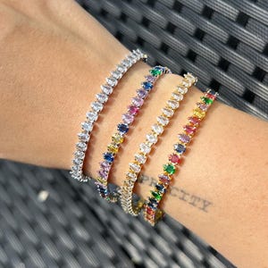 Könnte beinhalten: Vier farbenfrohe Tennisarmbänder werden an einem Handgelenk gezeigt. Die Armbänder verfügen über eine Vielzahl von Edelsteinen in weißen, Regenbogen- und Goldfassungen. Die Edelsteine sind in Marquiseform geschliffen.