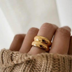 May include: A set of two stacked gold-toned rings with a smooth, rounded, and polished surface. The rings feature a thick, domed band design that sits flush against each other, creating a continuous, reflective metallic appearance on the finger.