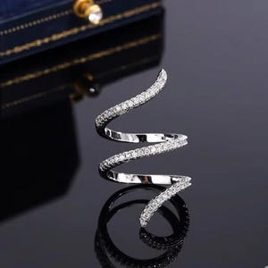 Può includere: Un anello a spirale argentato, incastonato con piccole pietre chiare scintillanti. Il design dell'anello si curva con grazia, creando un look moderno ed elegante. L'anello è presentato su una superficie nera riflettente.