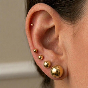 May include: Close-up of an ear with five gold ball earrings of varying sizes. The largest earring is a large, round stud, while the others decrease in size, creating a graduated effect. The earrings are shiny and reflect light.