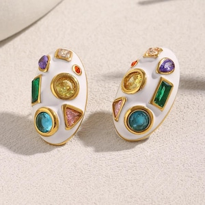 Peut inclure: Une paire de boucles d'oreilles ovales blanches avec des bordures dorées, chacune ornée de pierres précieuses colorées de différentes formes. Les boucles d'oreilles présentent un mélange de pierres vertes émeraude, bleues saphir et jaunes.