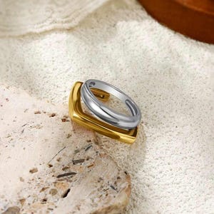 May include: A gold-coloured ring with a square base and a silver-coloured ring stacked on top. The rings are displayed on a textured, off-white surface. The rings have a modern, minimalist design.
