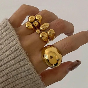 May include: Three gold rings are shown on a hand. One ring has a cluster of teardrop shapes, another has two connected rounded forms, and the third is a large, smooth oval. The rings have a shiny, reflective surface.