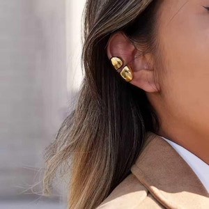 May include: Gold ear cuffs with a smooth, rounded design. The ear cuffs are worn on the ear. The image shows a close-up of the ear and the ear cuffs, with the person's hair and clothing in the background.