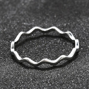 Può includere: Un anello color argento con un design ondulato distintivo. L'anello presenta un motivo continuo e fluido, creando un'estetica unica. L'anello è posto su uno sfondo scuro e strutturato.