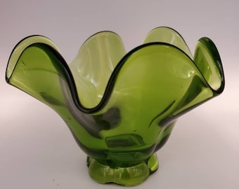 May include: A green glass bowl with a ruffled edge and a small, rounded base. The bowl is made of clear glass with a green tint.