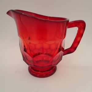 May include: A red glass pitcher with a faceted design and a handle. The pitcher is made of clear glass with a red tint.