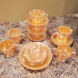 May include: A set of vintage, peach-colored, milk glass teacups and saucers. The set includes a creamer and sugar bowl. The cups and saucers have a delicate floral pattern.
