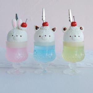 Puede incluir: Tres copas de postre decorativas, cada una llena de un líquido de diferente color y rematada con una confitería blanca con forma de animal y una cereza. Las copas son transparentes con un patrón decorativo.