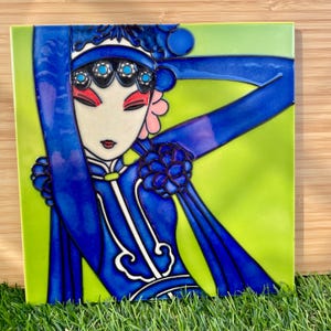 May include: A square ceramic tile featuring a stylised illustration of a figure in traditional opera attire. The design uses vibrant blue, lime green, and pink colours, showcasing a detailed headpiece with turquoise accents and a blue robe.