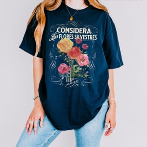 May include: A navy blue t-shirt with the text "CONSIDERA Las FLORES SILVESTRES" and a floral design in shades of yellow, red, and pink. The text "LUCAS 12:27" is below the floral design. The shirt is labeled "TRUE NAVY".