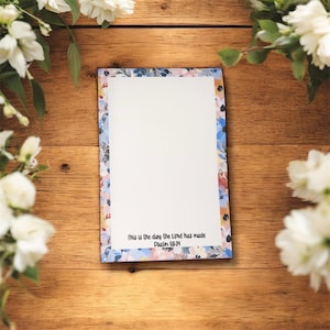 May include: A floral-bordered notepad with the text "This is the day the Lord has made Psalm 118:24" on a wooden surface. The border features a mix of blue, pink, and white flowers. White flowers and green leaves are arranged around the notepad.