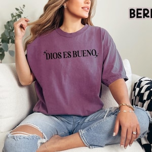 May include: Berry-colored t-shirt with the text "DIOS ES BUENO" in black. The shirt is a relaxed fit and is paired with blue jeans. The word "BERRY" is printed in the upper right corner. The person is sitting on a white couch.