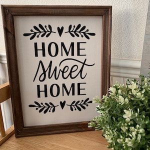 May include: A framed canvas sign with the text "Home Sweet Home" in black lettering. The sign is decorated with small black floral designs and hearts. The sign is sitting on a wooden surface.