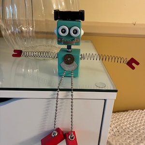 May include: A handmade teal robot sculpture with a black head, googly eyes, and spring arms. The robot has a coin on its chest and red wooden feet hanging from a chain. A unique, whimsical art piece.