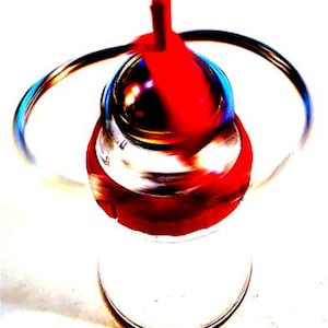 May include: A clear glass bottle with a red and silver stopper. The stopper has a red top and a silver base. A silver wire handle arches over the top of the bottle. The bottle is on a white surface.