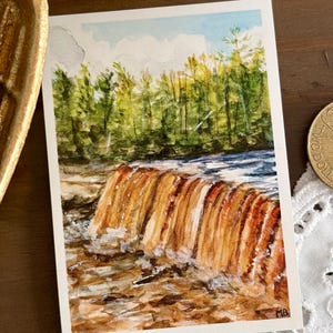 May include: A watercolor painting of a waterfall cascading over rocks. The water is a mix of white and blue, with the waterfall itself a warm brown. Green trees and a blue sky complete the landscape. The painting is on a white rectangular card.