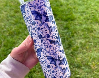 May include: A white and blue stainless steel tumbler with a floral pattern and bats. The tumbler has a straw and is being held in a hand.