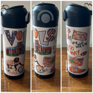 May include: A white stainless steel tumbler with a black lid and a variety of orange and white graphics. The graphics include the words "Go Vols!" and "Big Orange", a football helmet, a stadium, and a building. The tumbler is decorated with a black and orange striped pattern.