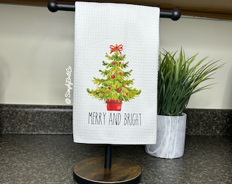 Peut inclure: Essuie-mains blanc avec un motif de sapin de Noël, un nœud rouge et l'inscription « MERRY AND BRIGHT ». Le torchon est présenté sur un support en métal noir avec une base en bois. Une petite plante en pot est à l'arrière-plan.
