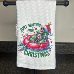 May include: White kitchen towel with a skeleton wearing a Santa hat and a Christmas tree float. The text reads "Just Waiting For Christmas".
