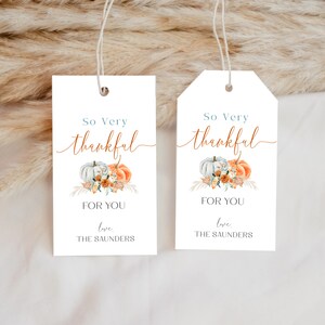 以下が含まれることがあります： かぼちゃ、花、秋の紅葉の水彩画が描かれた白いギフトタグ2枚。タグには「So Very Thankful」と「For You」の文字が書かれている。タグの下部には「Love, The Saunders」と書いてある。