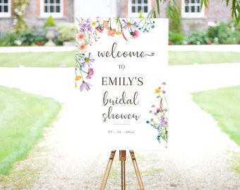 May include: A white welcome sign with a floral border and the text "Welcome to Emily's Bridal Shower 05.26.20XX". The sign is on a wooden easel in front of a green lawn and a brick building.