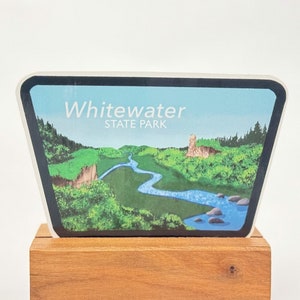 May include: A wooden stand with a colorful sticker featuring a scenic view of a river winding through a forest. The sticker reads "Whitewater State Park".
